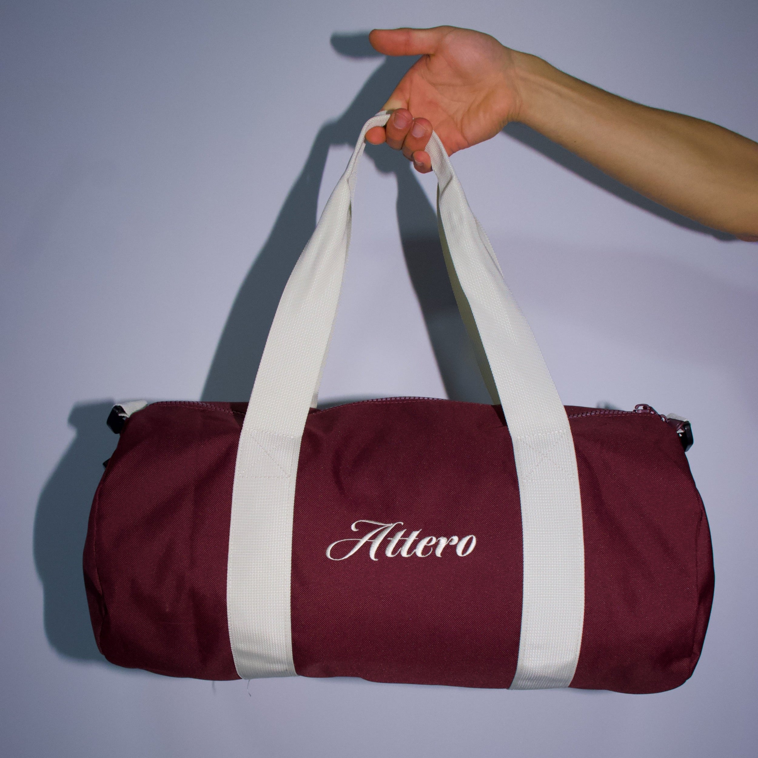 Burgundy ROMA duffel bag by Attero with white straps and embroidered logo, held by hand against a light background.